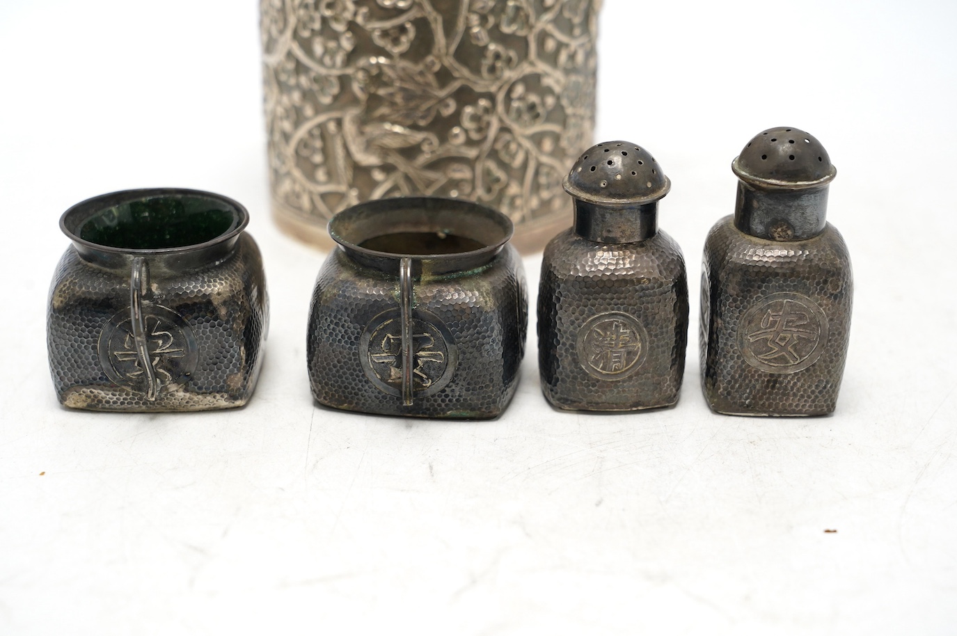 A Chinese embossed white metal cylindrical jar and cover, 11.2cm, and a similar white metal four piece cruet set. Condition - poor to fair